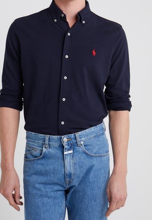 Mężczyzna w granatowej koszuli na guziki z czerwonym wyszywanym logo i jasnoniebieskich dżinsach, z podwiniętymi rękawami i rękami swobodnie opuszczonymi wzdłuż ciała.