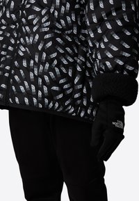 Svart vattert jakke med hvitt geometrisk mønster, quiltet design og myke svarte mansjetter. Komplett med en svart hanske med en logo.