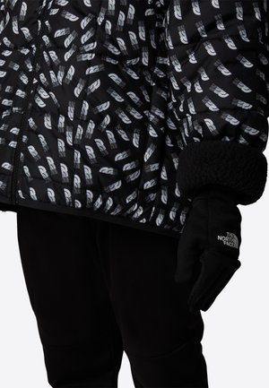 Czarna pikowana kurtka puchowa z białym geometrycznym wzorem, pikowanym designem i miękkimi czarnymi mankietami. Towarzyszy jej czarna rękawiczka z logo.