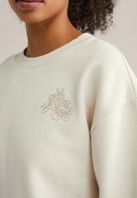 Nahaufnahme einer Person, die ein cremefarbenes Sweatshirt mit einem zarten, gestickten Blumenmuster auf der Brust nahe der Schulter trägt.