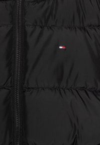 Czarna pikowana kurtka puchowa z zamkiem błyskawicznym z przodu i małym haftowanym logo w kolorach czerwonym, białym i niebieskim na lewej piersi. Gładki, błyszczący materiał.