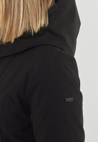 Giacca nera con cappuccio e una texture liscia, dotata di una toppa con logo sulla manica sinistra e una silhouette aderente.