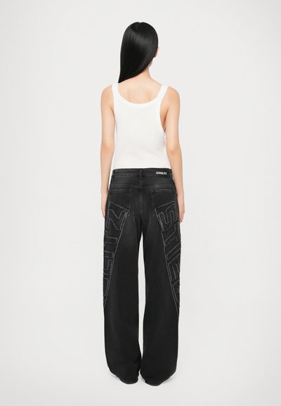 Black wide-leg jeans with distressed stitching and “STIGLITZ” embroidered on the sides. Paired with a plain white tank top.
