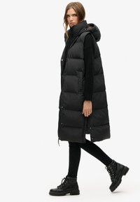 Schwarze Daunenweste mit Kapuze, mit einem gesteppten Design und Druckknöpfen. Kombiniert mit schwarzen engen Hosen und robusten schwarzen Schnürstiefeln.