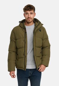 Mand iført en olivengrøn pufferjakke med hætte, lysegrå struktureret sweater og blå jeans, stående foran en ensfarvet hvid baggrund.