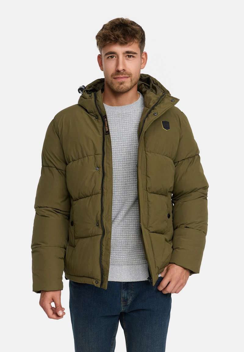 Mand iført en olivengrøn pufferjakke med hætte, lysegrå struktureret sweater og blå jeans, stående foran en ensfarvet hvid baggrund.