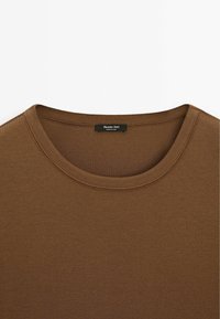 T-shirt marron à col rond avec une couture visible au niveau du col et une étiquette noire cousue à l'intérieur de l'encolure.