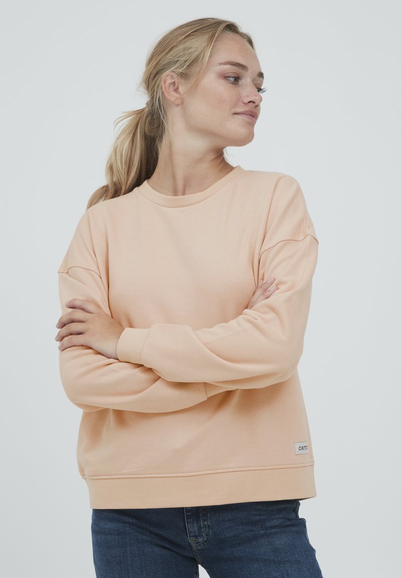 Lys fersken sweatshirt med rund hals, lange ermer og ribbestrikkede mansjetter. Har en liten merkelapp med logo ved nederkant, glatt tekstur.