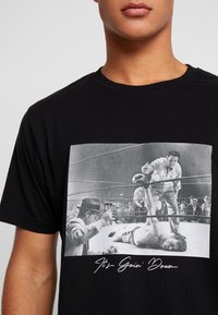 T-shirt noir en coton avec un graphique de scène de boxe vintage en niveaux de gris, avec le texte blanc "Ça va chauffer" en dessous de l'illustration.