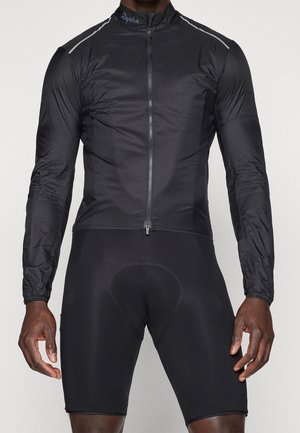 Athlete wearing black long-sleeve cycling jacket with front zipper and black padded cycling shorts against light background.