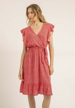Femme portant une robe rouge mi-longue à motifs floraux blancs, col en V, manches volantes et ceinture nouée à la taille, debout devant un fond uni.