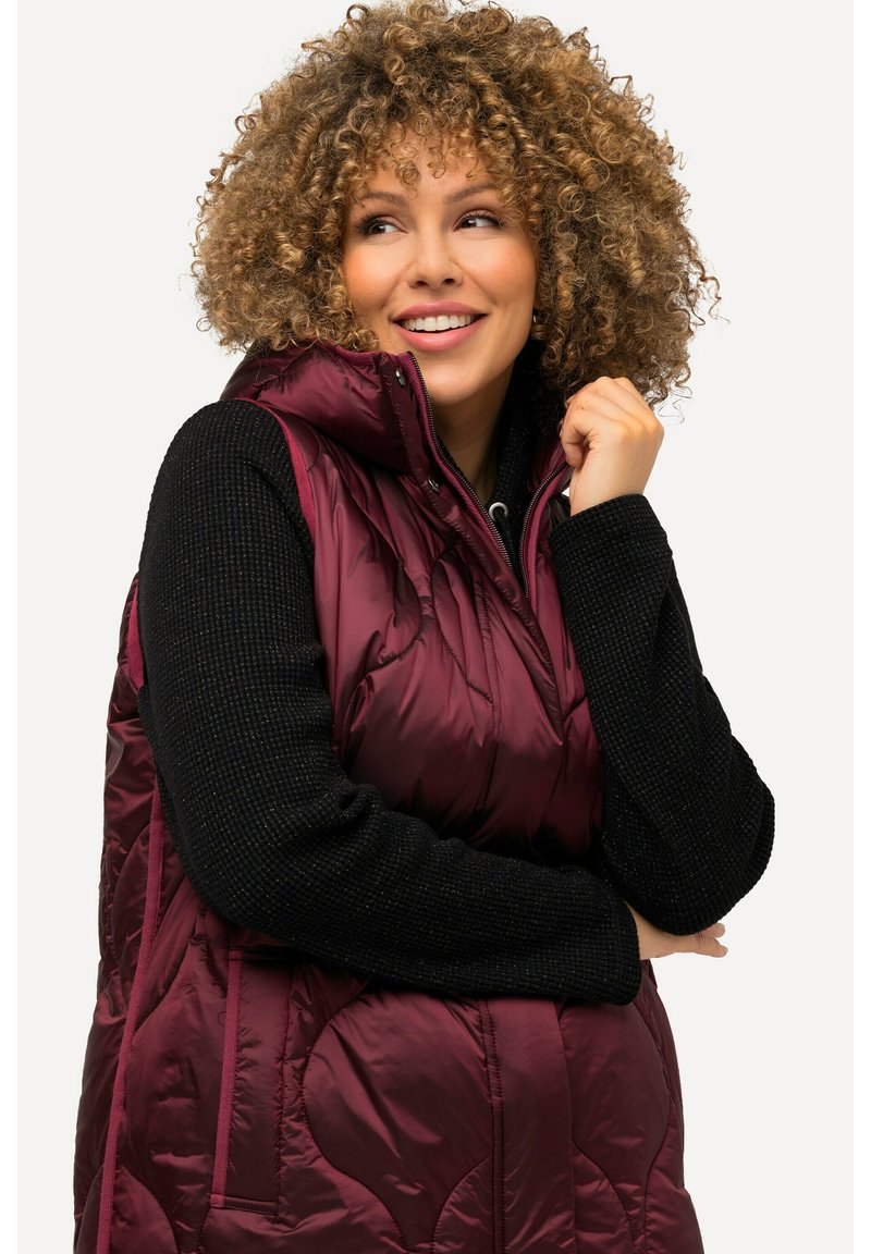 Bordeauxkleurige puffer vest met hoge kraag, glanzende afwerking en gestikt patroon, gedragen over een gestructureerde zwarte trui. Trekkoord bij de hals.