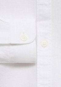 Valkoinen paitakangas, jossa on teksturoidun näköinen pinta, varustettuna taitetulla hihansulla ja kahdella kermavärisellä napilla, joissa on neljä reikää kumpikin.