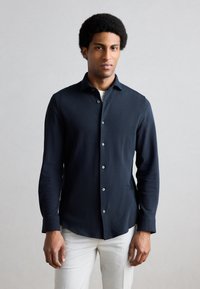 Granatowa koszula z długim rękawem, zapinana na guziki z przodu, o dopasowanym kroju i subtelnej fakturze; noszona z jasnymi spodniami.