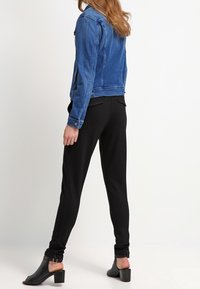 Žena oblečená v modré džínové bundě, černých přiléhavých kalhotách a černých kožených kotníkových botách, stojící na bílém pozadí.