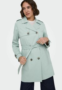 Femme attachant une ceinture sur un trench-coat bleu clair à double boutonnage avec des poignets à boutons, debout devant un fond blanc uni.
