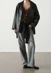 Svart läderjacka över en brun topp med spetskant, gråvida jeans med vida ben och en svart tröja knuten i midjan. Svarta skor kompletterar looken.