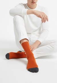 Oranžna nogavica s temnosivimi deli na prstih in peti, izdelana iz teksturiranega materiala. Oseba jo nosi skupaj z belo majico z dolgimi rokavi in hlačami.
