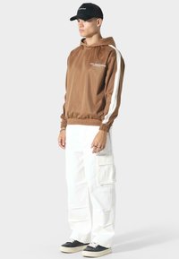 Bruine satijnen hoodie met witte zijstrepen, elastische manchetten en zoom. Gecombineerd met witte cargobroeken met meerdere zakken en zwarte sneakers.