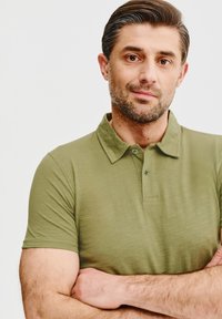 Oliwkowa koszulka polo z kołnierzykiem, krótkimi rękawami i zapięciem na dwa guziki. Miękka tekstura z delikatnym połyskiem. Prosty design bez wzorów.