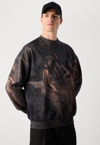 Ung mand iført en sort kasket og en mørk oversized sweatshirt med et vintage-inspireret maleri af kvinder i kjoler på fronten og ærmerne.
