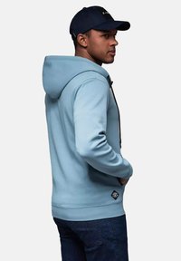 Lyseblå hættetrøje med snoretræk, blødt stof og sidelommer. Båret over blå denim, med et logo-patch nær kanten.