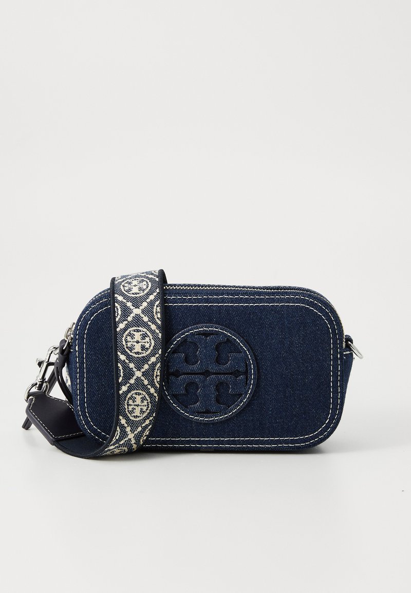 Bolsa de ombro em denim com costura branca e um logotipo. Apresenta uma alça ajustável com padrão e um fecho com zíper. A cor principal é azul-escuro.