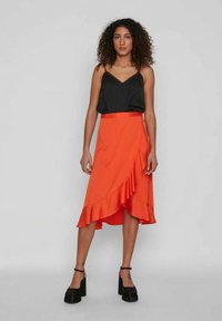 Oranža wrap stila svārki ar volāniem apakšmalā, kas saskaņoti ar melnu krekliņu ar plānām siksnām. Modelis valkā masīvus melnus platformas papēžus.