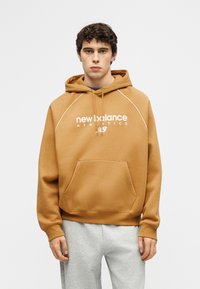 Νεαρός άνδρας που φοράει μπεζ φούτερ New Balance με λευκό λογότυπο και ανοιχτό γκρι παντελόνι φόρμας, στέκεται μπροστά σε ένα απλό ανοιχτό φόντο.