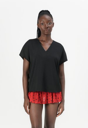 Vrouw met gevlochten haar die een loszittende zwarte V-hals top draagt over een rode rok met zwarte bolletjes, staand tegen een eenvoudige witte achtergrond.