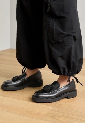 Mocassins en cuir noir avec un détail de gland, dotés d'une semelle en caoutchouc épaisse et de coutures texturées, associés à un pantalon noir ample.