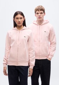 Ανοιχτό ροζ φούτερ με φερμουάρ και κουκούλα, κατασκευασμένα από απαλό ύφασμα, με τσέπες τύπου καγκουρό και το λογότυπο της Lacoste στην αριστερή πλευρά του στήθους. Φοριούνται από δύο μοντέλα.