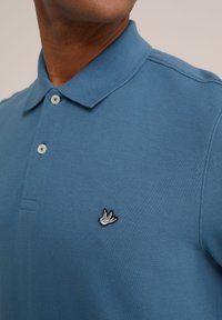 Modra polo majica iz teksturiranega blaga, z izvezeno črno-belo ptico na prsih in svetlimi gumbi na ovratniku.