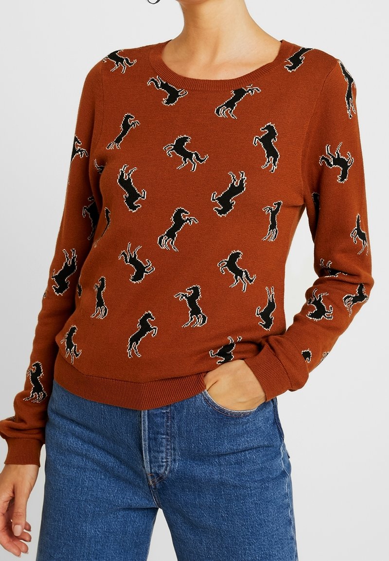 Sweater en laine marron avec des motifs de chevaux noirs, encolure ronde et poignets côtelés. Associé avec un jean en denim bleu.