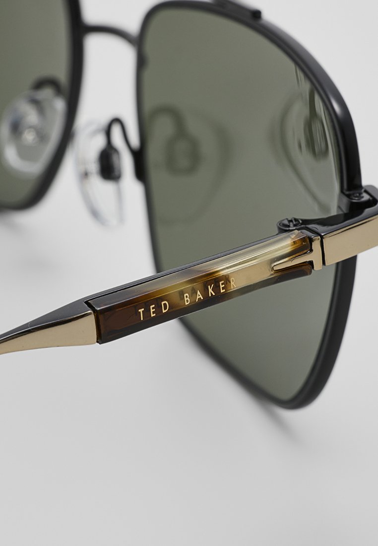 Sonnenbrille mit dunkelgrünen Gläsern und einem schwarz-goldenen Rahmen, mit dem "Ted Baker"-Logo in goldener Schrift auf dem Bügel.