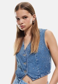 Chaleco de mezclilla en azul claro con un diseño cropped, que presenta botones y adornos metálicos en forma de estrella. Textura suave con corte sin mangas.