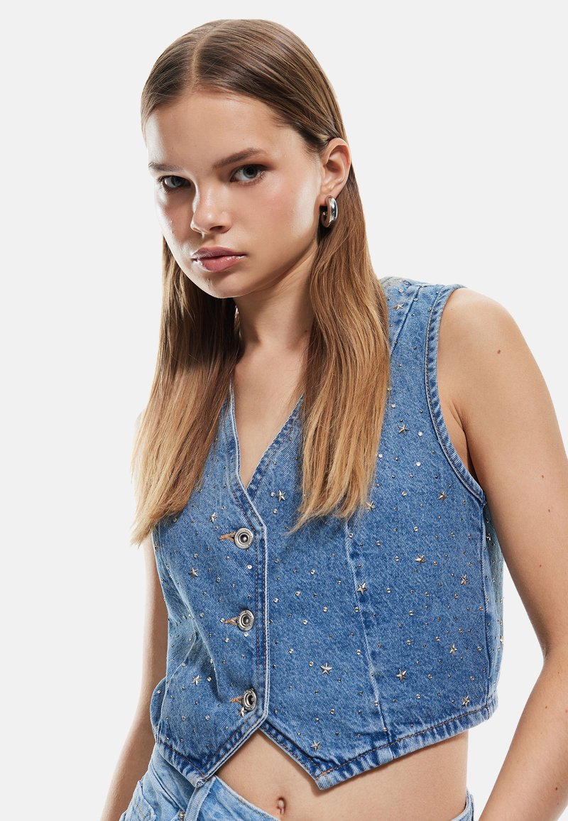 Chaleco de mezclilla en azul claro con un diseño cropped, que presenta botones y adornos metálicos en forma de estrella. Textura suave con corte sin mangas.