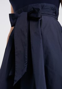 Mörkblå klänning med en figursydd överdel och en stor rosett i midjan samt en flödande A-linje-skjol, tillverkad av slät, texturerad tyg.