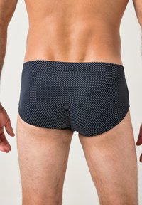Torso e gambe maschili che indossano un costume da bagno scuro con piccoli pois bianchi, mostrati di spalle su uno sfondo semplice.