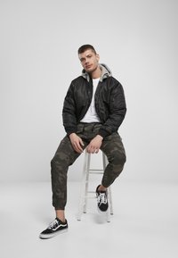 Jachetă bomber neagră cu glugă gri, tricou alb, pantaloni cargo cu camuflaj și adidași negri, așezat pe un scaun alb în fața unui fundal gri.