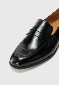 Mocassin en cuir verni noir avec une finition lisse, un bout arrondi et une sangle décorative au-dessus. Détail de couture subtil.