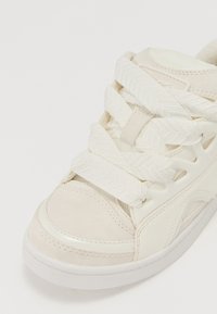 Adidași albi cu partea superioară din piele netedă, accente texturate și șireturi groase. Au un vârf rotunjit și laterale perforate pentru ventilație.