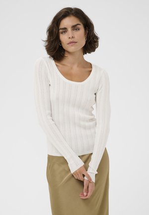 Žena s krátkymi hnedými vlasmi, oblečená v bielom pletenom tričku s dlhými rukávmi a béžovou sukňou, stojaca so zloženými rukami pred sebou.