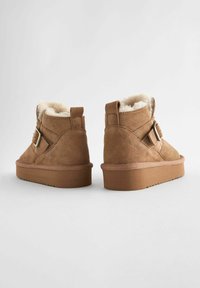 Stivaletti alla caviglia in suede marrone con rivestimento in pelliccia sintetica, cinturino regolabile e suole in gomma spessa. Caratterizzati da una punta tonda e un tacco texturizzato.