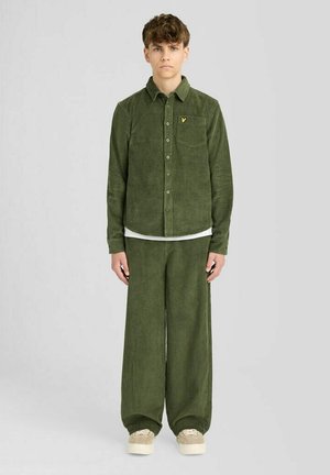 Jong persoon staat, draagt een olijfgroen corduroy overhemd en wijde broek met beige sneakers op een effen lichte achtergrond.