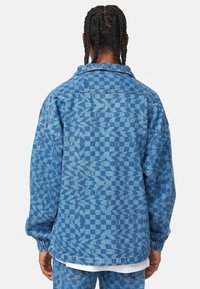 Veste en denim bicolore bleu avec des motifs géométriques. Coupe oversize, manches longues et poignets élastiques. Texture subtile avec un col.