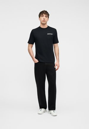 Νέος άνδρας στέκεται μπροστά σε απλό φόντο, φορώντας μαύρο μπλουζάκι Napapijri, μαύρο παντελόνι και λευκά αθλητικά παπούτσια, με το ένα χέρι στην τσέπη.