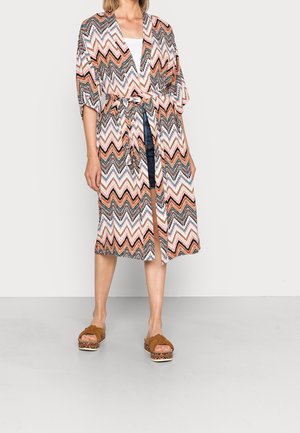 Femme portant une robe mi-longue aux genoux avec des motifs en zigzag noir, blanc, rose et marron, nouée à la taille, assortie à des sandales à plateforme marron.