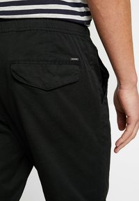 Czarne bawełniane spodnie z tylną kieszenią z zaokrąglonym klapkiem i małym metalowym akcentem w postaci logo. Luźny krój z elastycznym paskiem w talii.