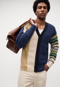 Strikcardigan i marineblå, beige og grønne farver. Har V-hals, knaplukning og mønstrede ærmer. Brun lædertaske båret over skulderen.